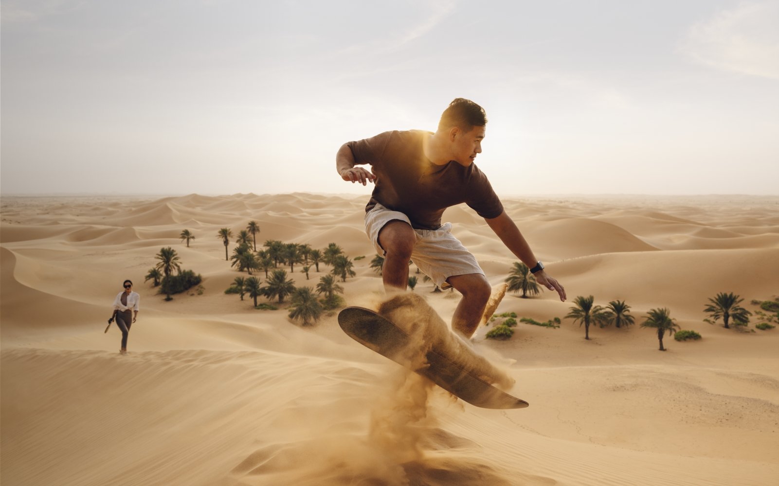 Man sandboarding on the dunes of Abu Dhabi with his wife in the background