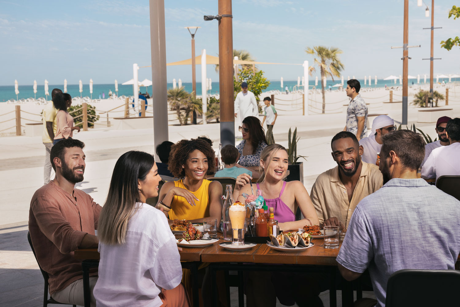 Group of friends dining and laughing at a beachfront restaurant on a sunny day.
