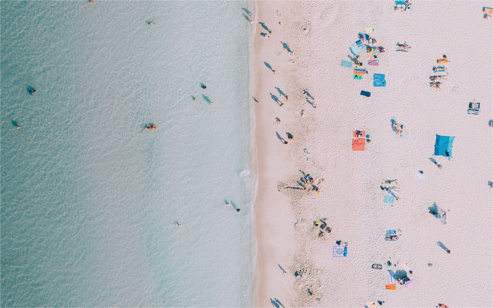Aerial view of beachgoers on sand and swimmers in clear turquoise water.