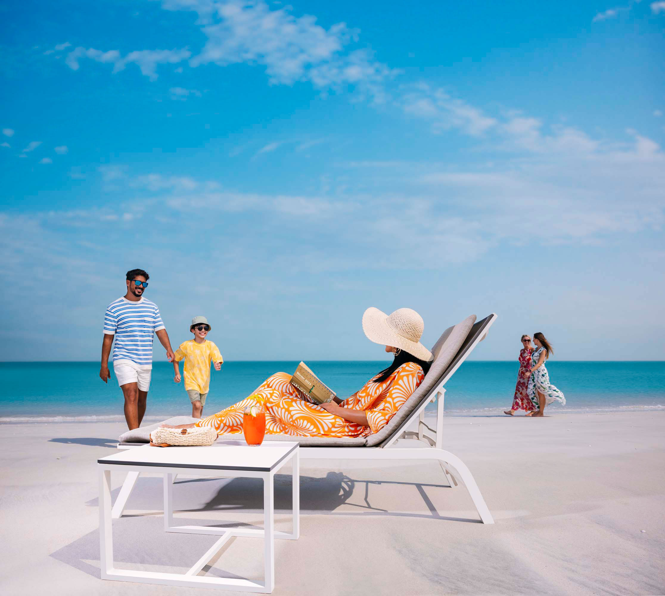 Woman reading on beach lounger while family and friends enjoy a walk by the turquoise sea.