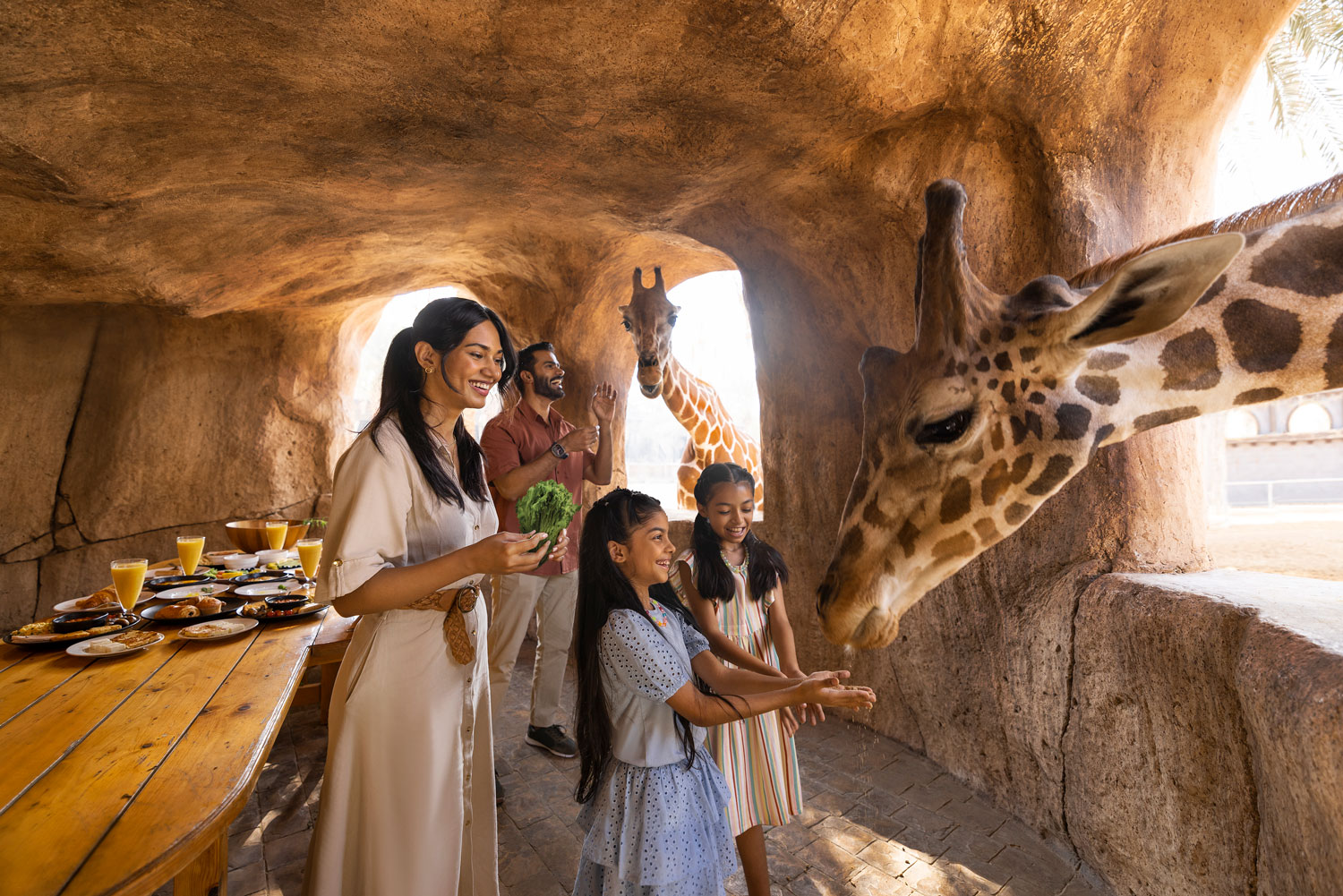 Family feeding giraffes in a shaded zoo dining area with breakfast spread on wooden table