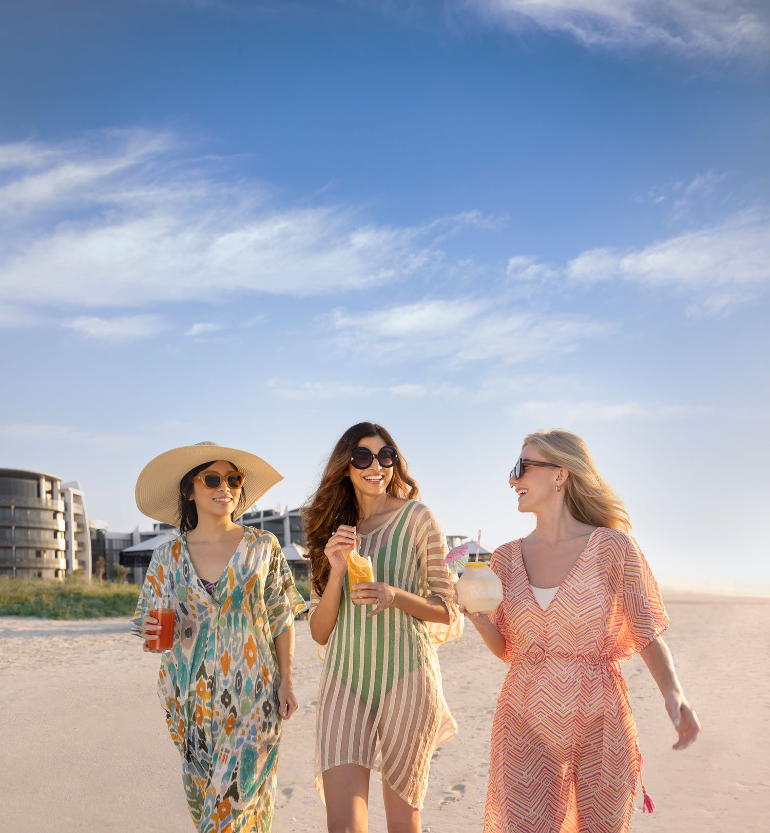Three women in colorful beachwear enjoying fresh drinks and walking along a sunny beach in Abu Dhabi