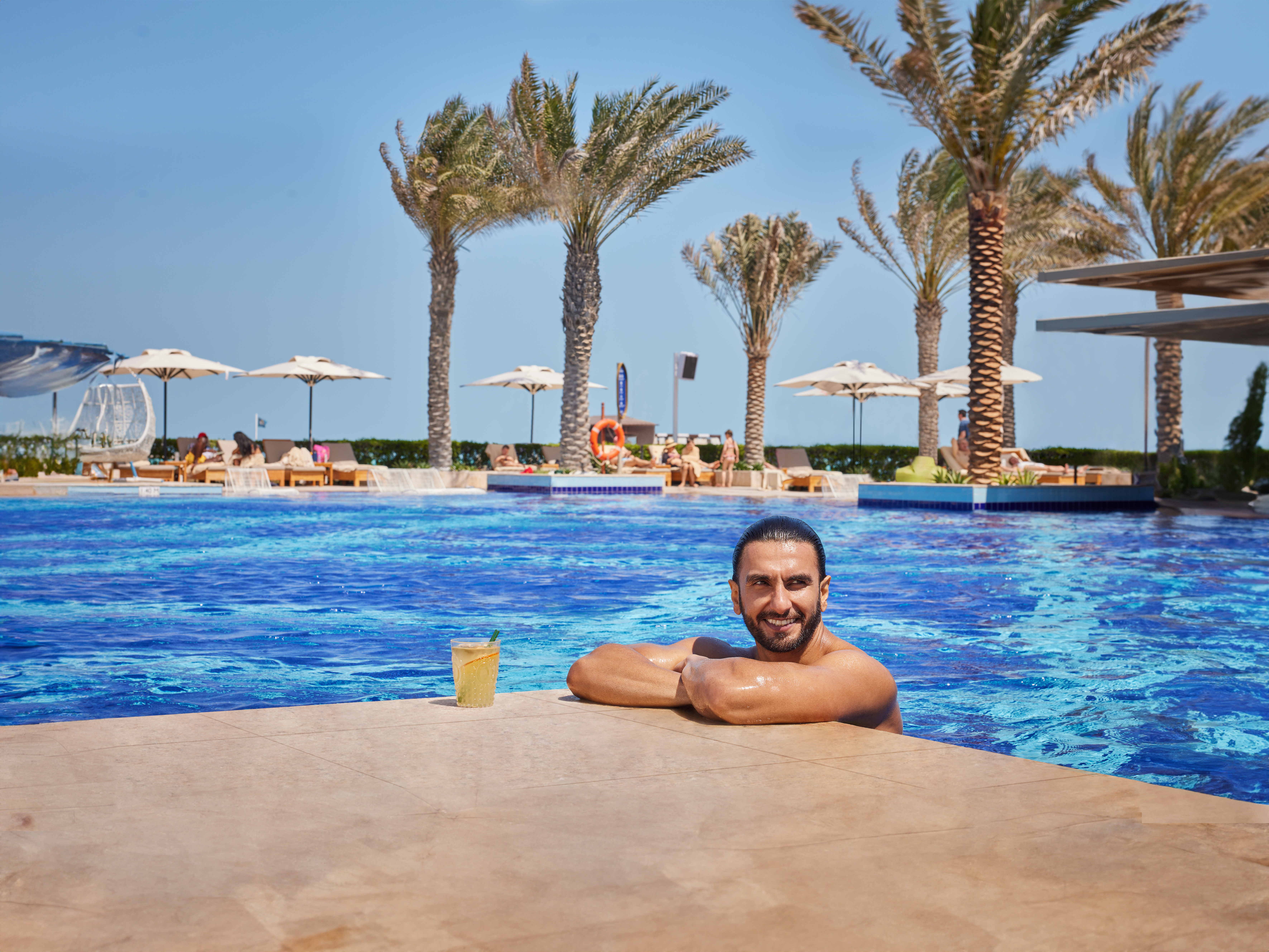 Man relaxing in a luxury pool with a drink, surrounded by palm trees and sun loungers in the background