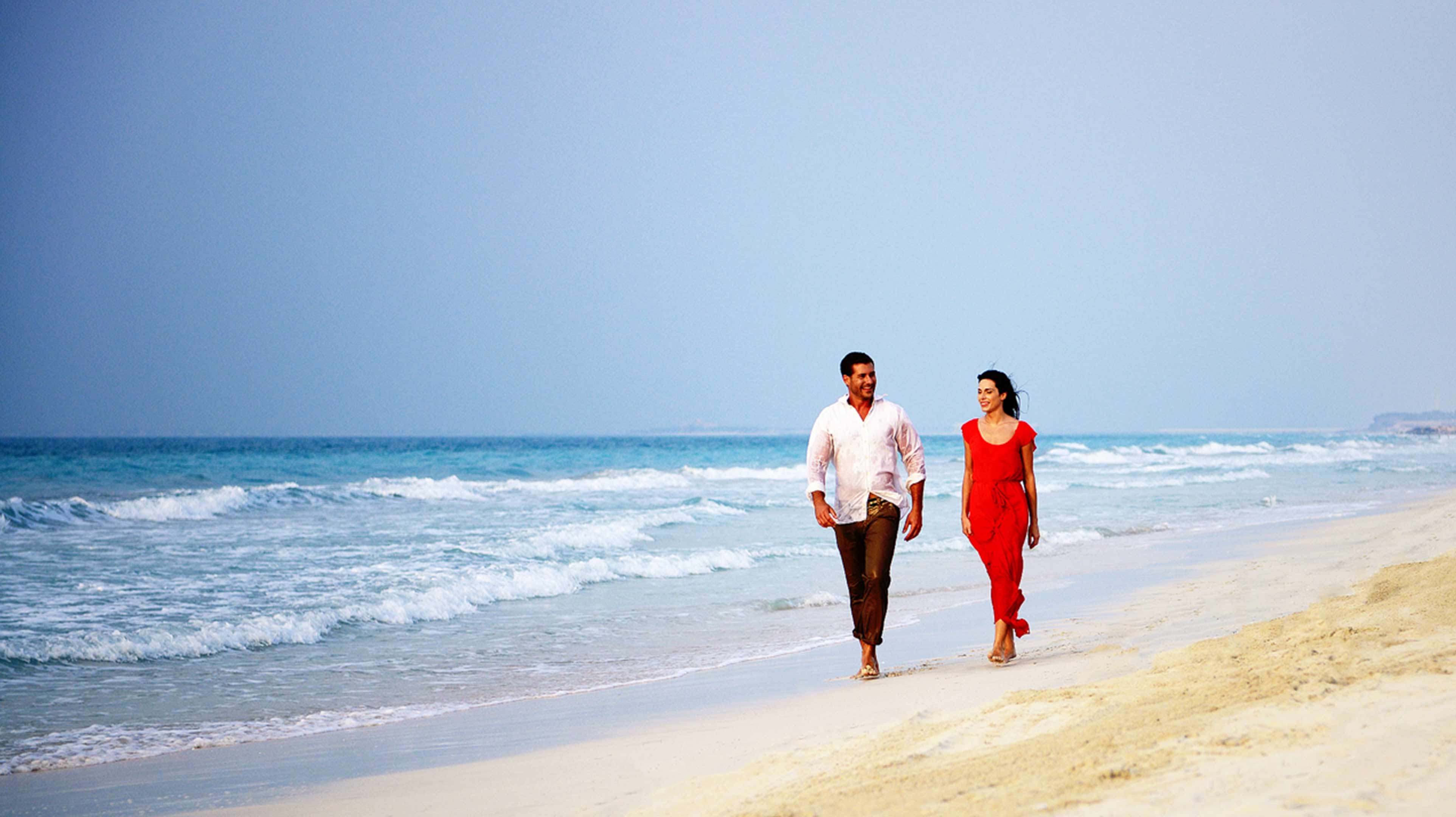 Una donna in un vestito rosso e un uomo passeggiano in spiaggia.