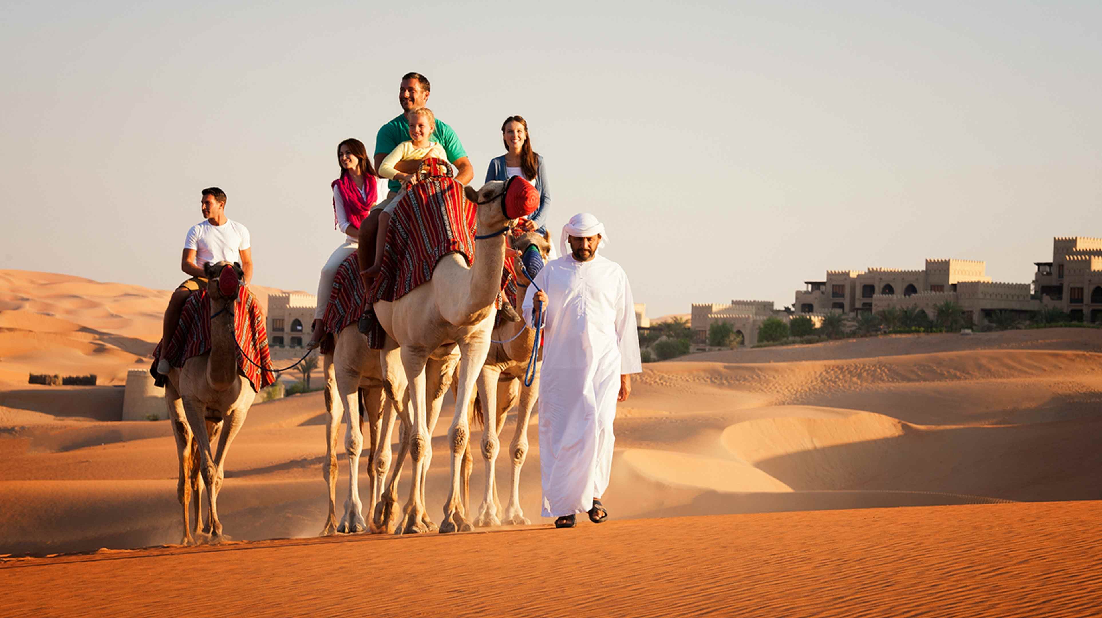 Western family riding camels outside the Qasr Al Sarab Desert Resort by Anantara with an Emirati guide