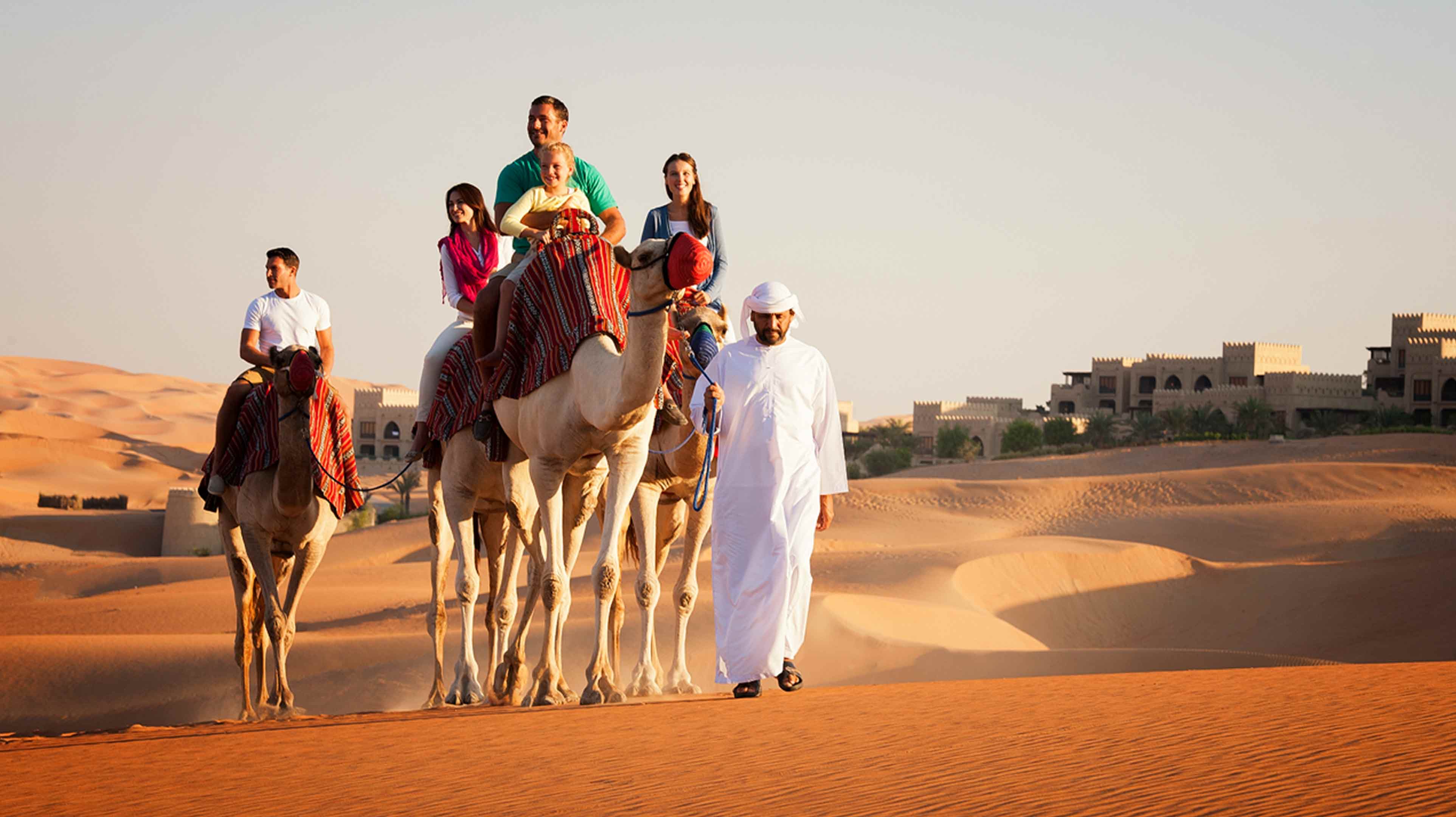 Piccolo gruppo di turisti sorridente lascia l'Anantara Qasr al Sarab Resort a dorso di cammello accompagnato da un uomo a piedi in abiti tradizionali