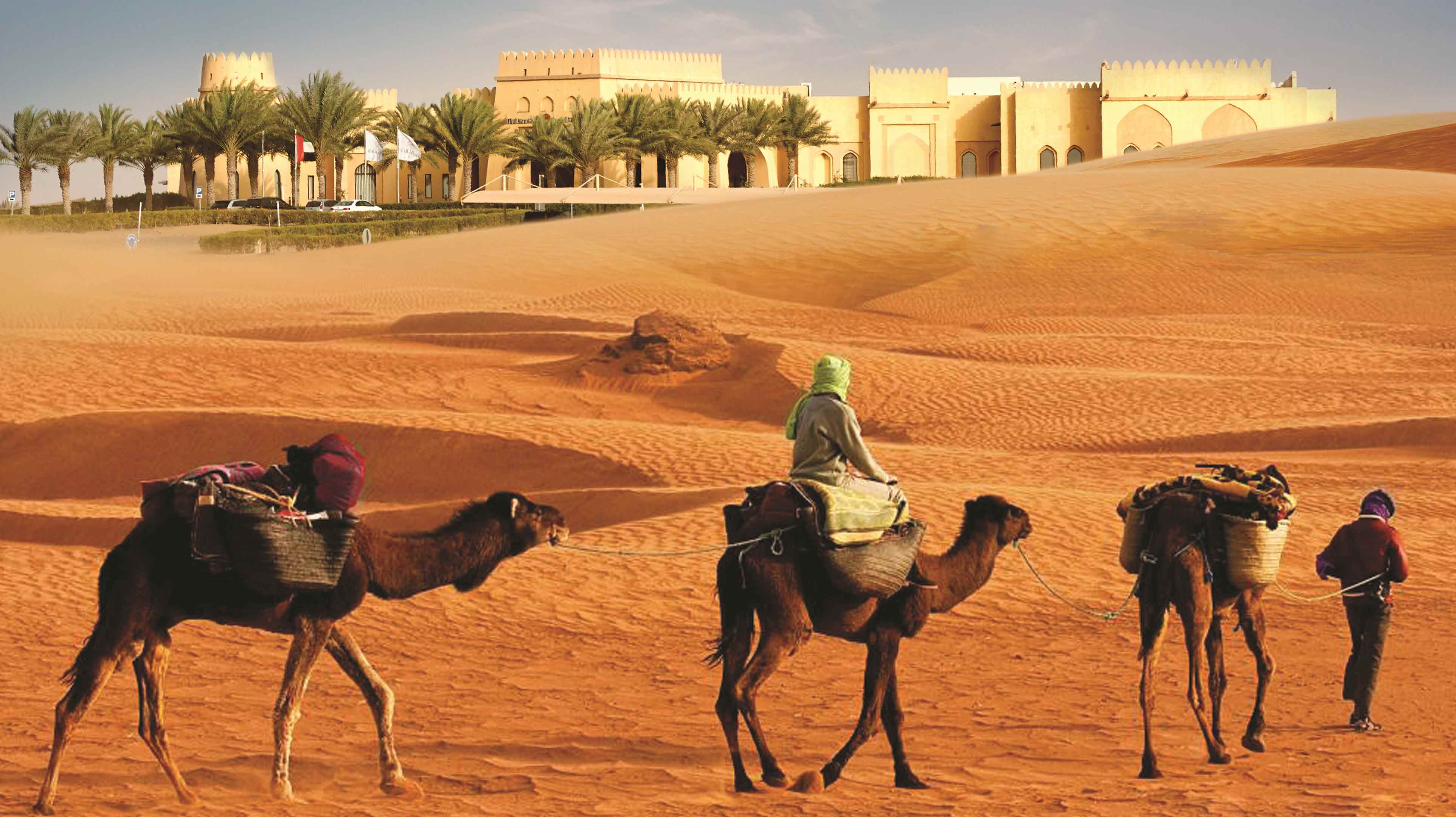 Tre cammelli in fila guidati da un giovane a piedi e da un uomo in abiti tradizionali seduto sul cammello di mezzo si muovono nel deserto Rub' al Khali di Abu Dhabi in direzione del Qasr Al Sarab Desert Resort by Anantara