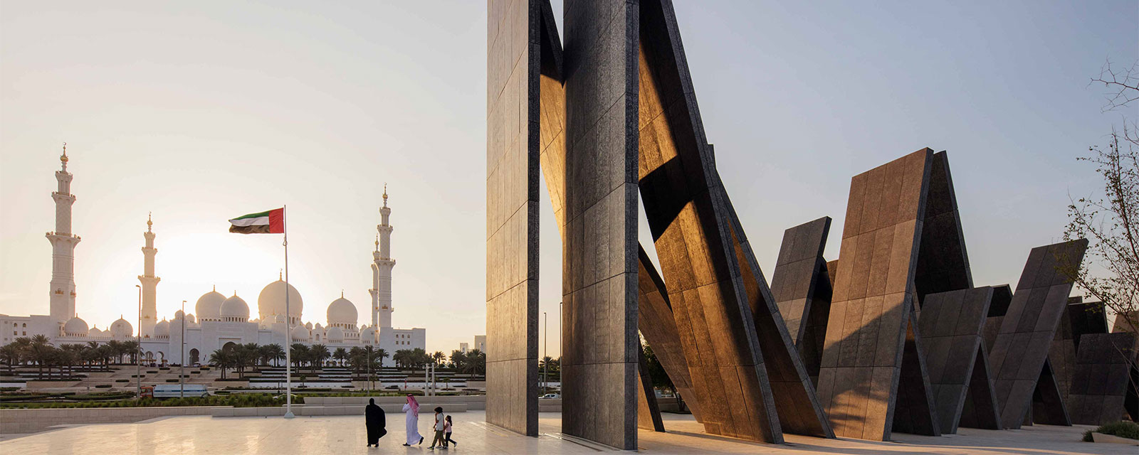  Devant le Wahat Al Karama, un couple se promène paisiblement, admirant la beauté de la Mosquée Cheik Zayed au coucher de soleil.