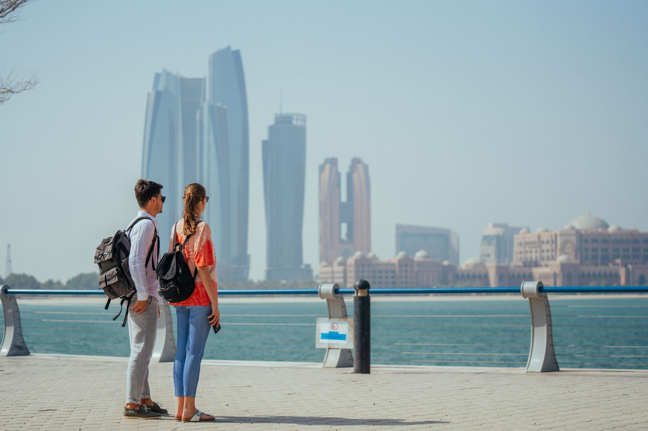Sur la Corniche d’Abu Dhabi, un couple contemple l'horizon scintillant, parfaite symbiose entre mer turquoise, nature apaisante et modernité urbaine.
