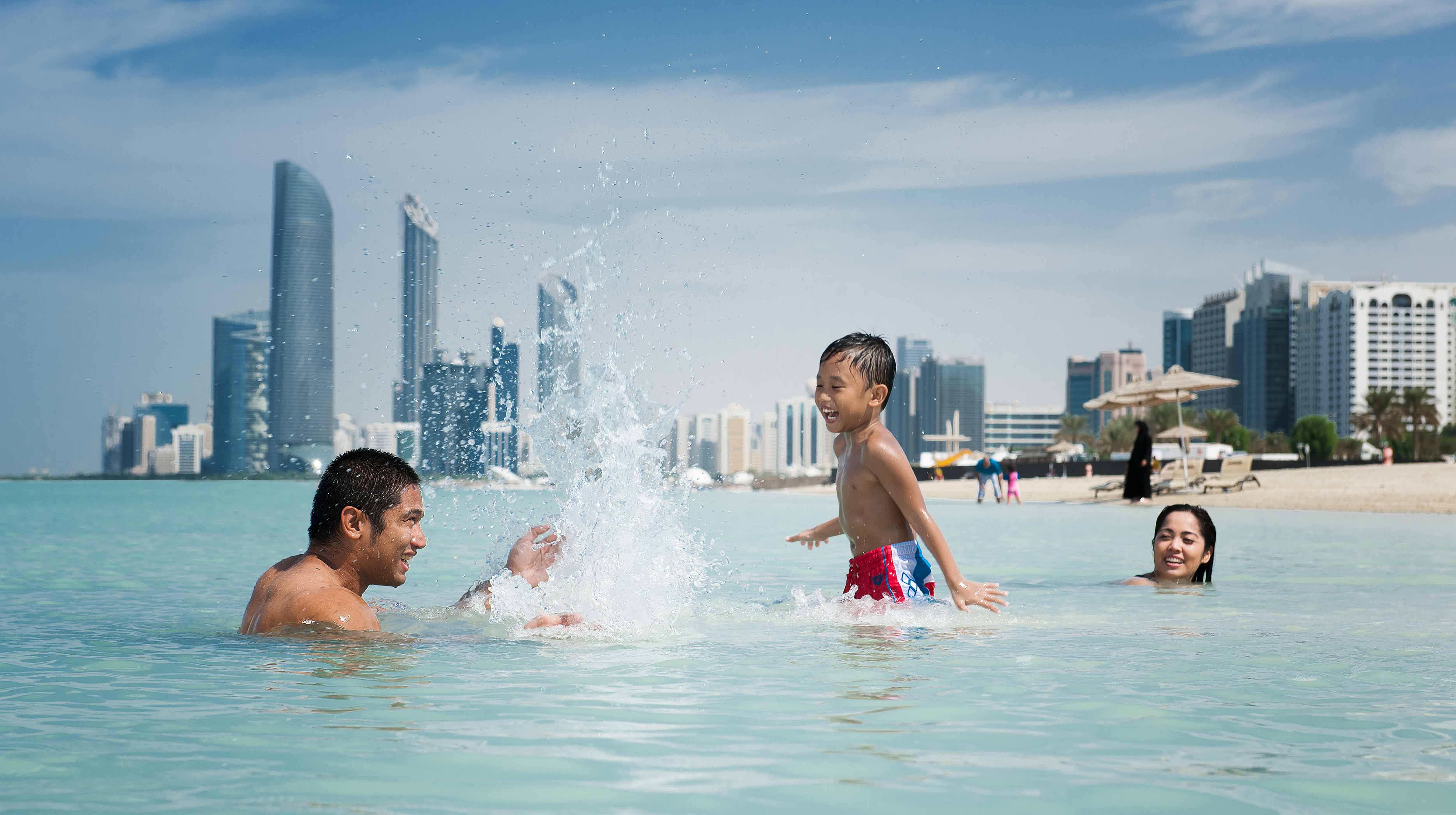 Una famiglia con papà, mamma e bambino si diverte a giocare nelle acque della spiaggia Corniche Beach con lo sfondo dei grattacieli di Abu Dhabi