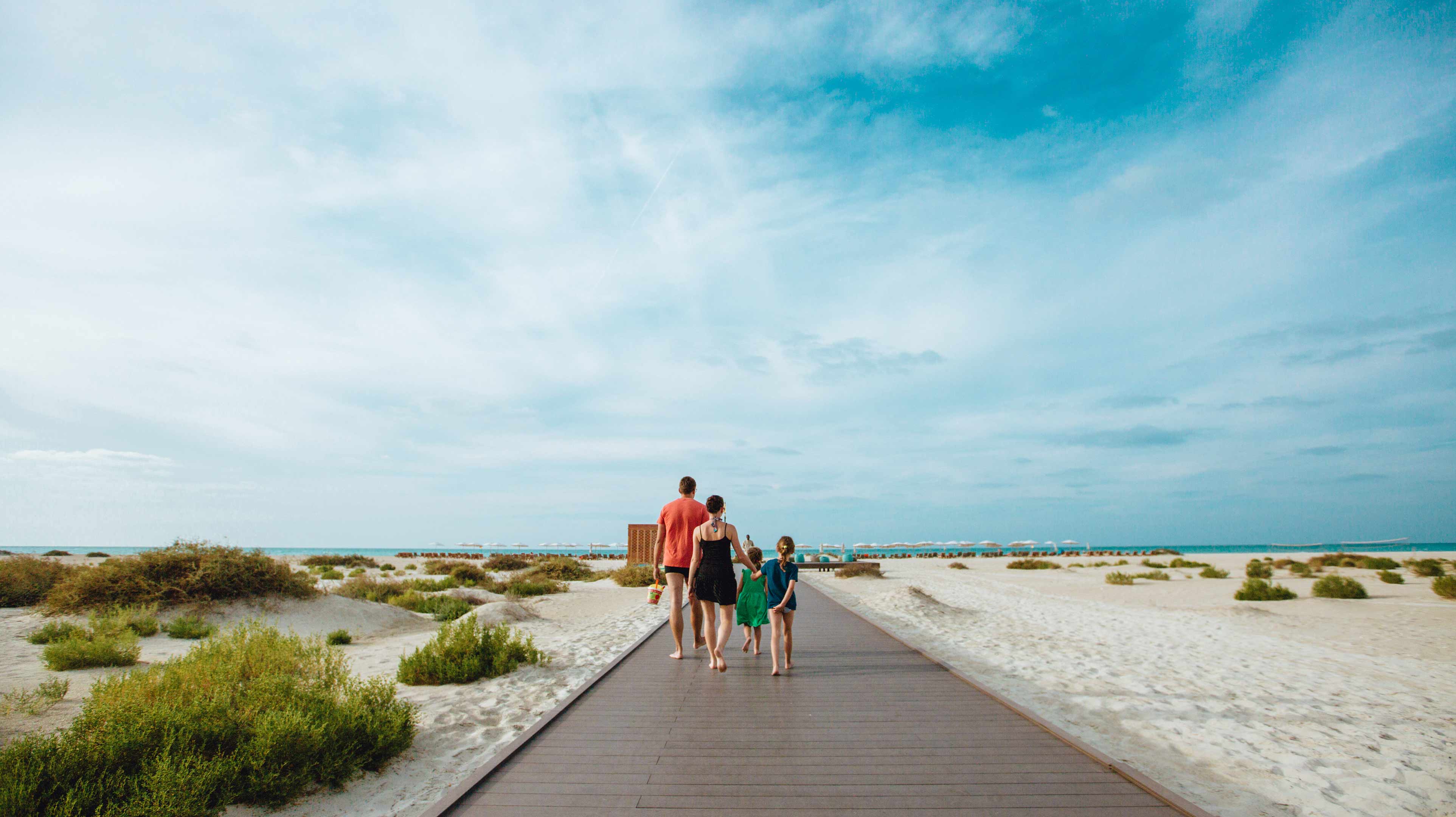 Una famiglia con mamma, papà e due bambini cammina verso il mare su una passerella di legno che attraversa la sabbia bianca e i cespugli di vegetazione della Kai Beach sull'isola di Saadiyat ad Abu Dhabi 