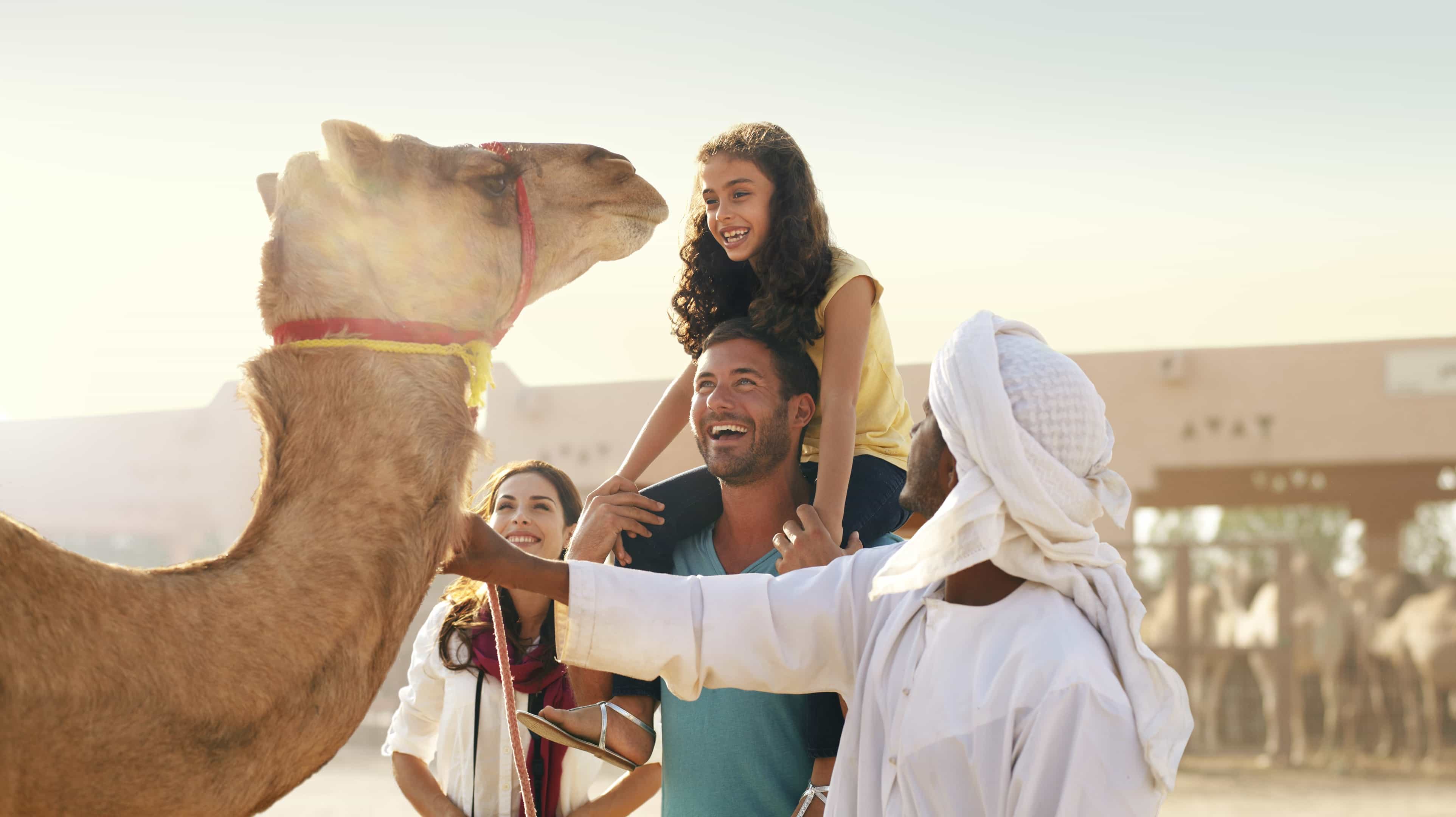 Famiglia di turisti sorridenti davanti a un cammello condotto da un uomo in abiti tradizionali al caratteristico mercato dei cammelli Al Ain Camel Market di Abu Dhabi
