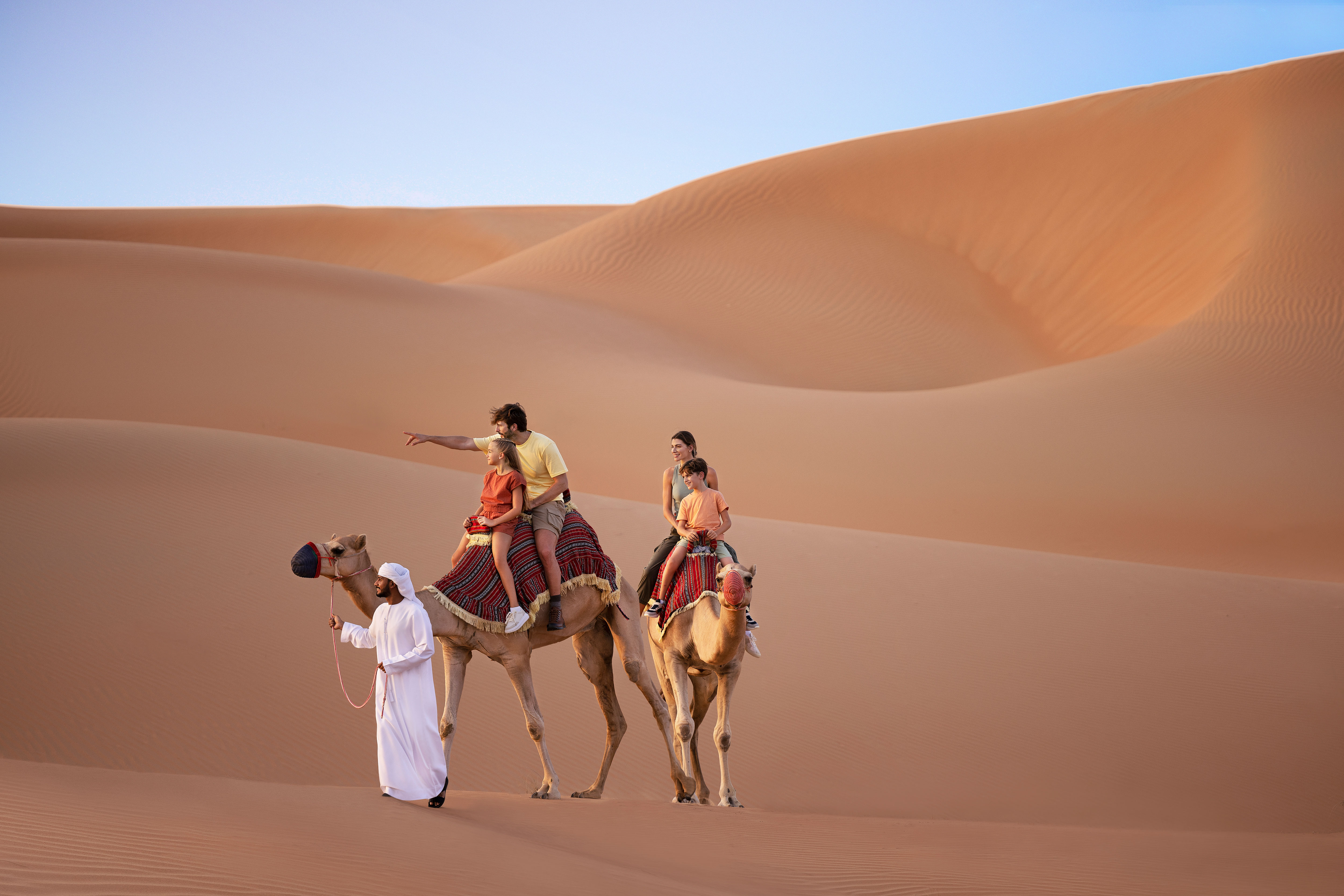 مرشد سياحي يقود جملين يركبهما عائلة من أربع أفراد وسط الكثبان الرملية في أبوظبي