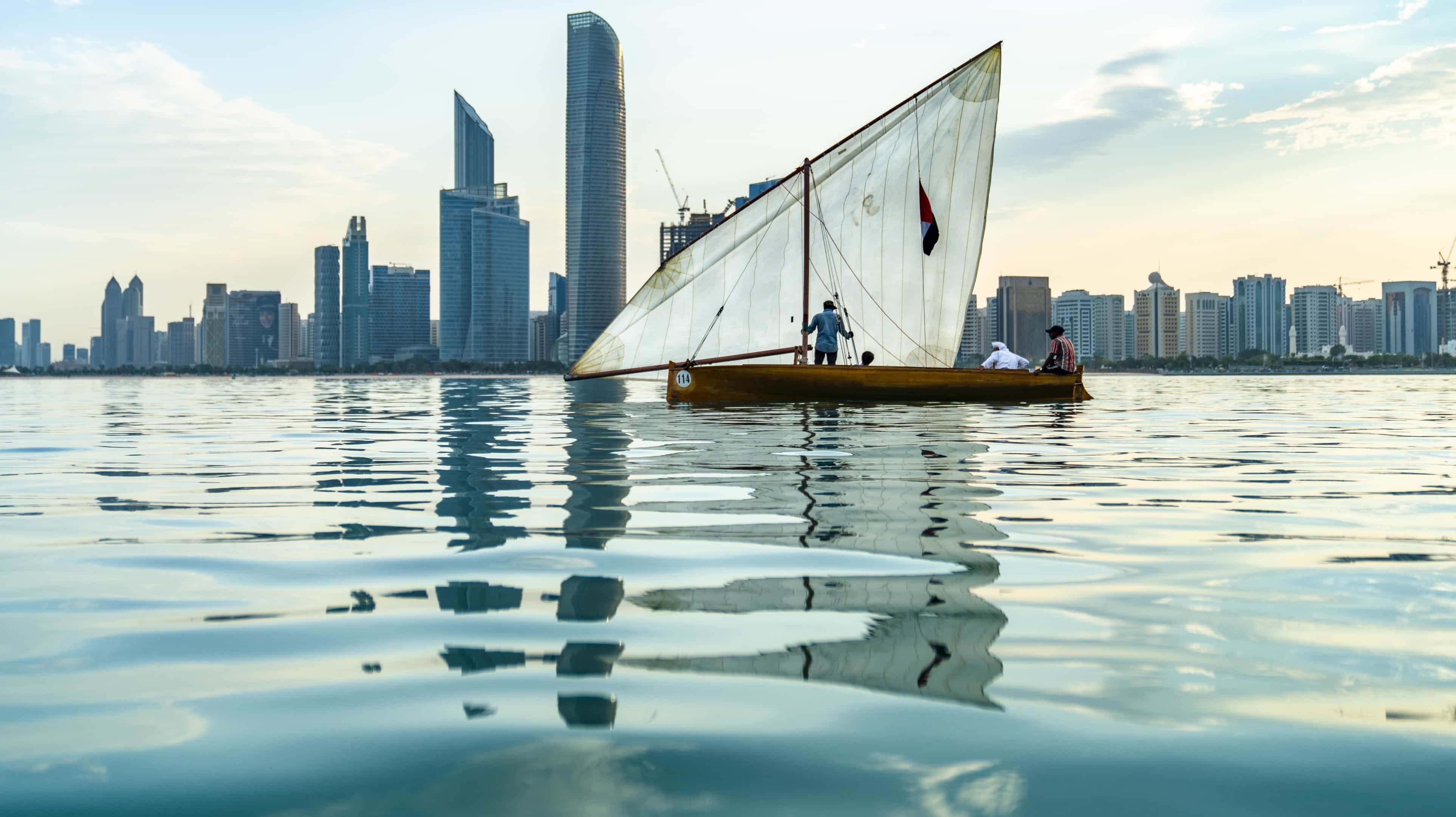 Barca a vela con tre persone sull'acqua. Si vede lo skyline di Abu Dhabi sullo sfondo.