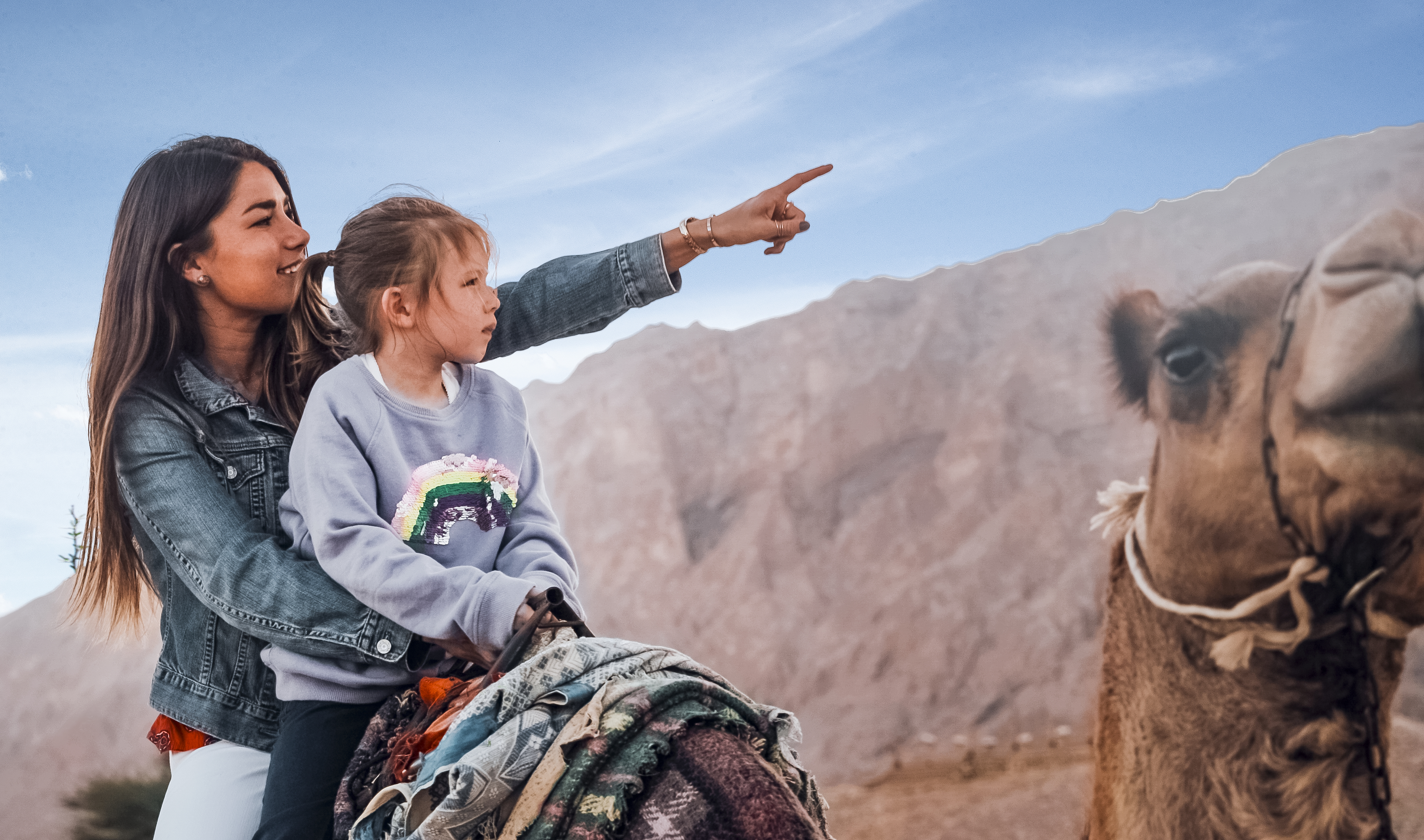 Woman and girl on a camel's back pointing at Jebel Hafit
