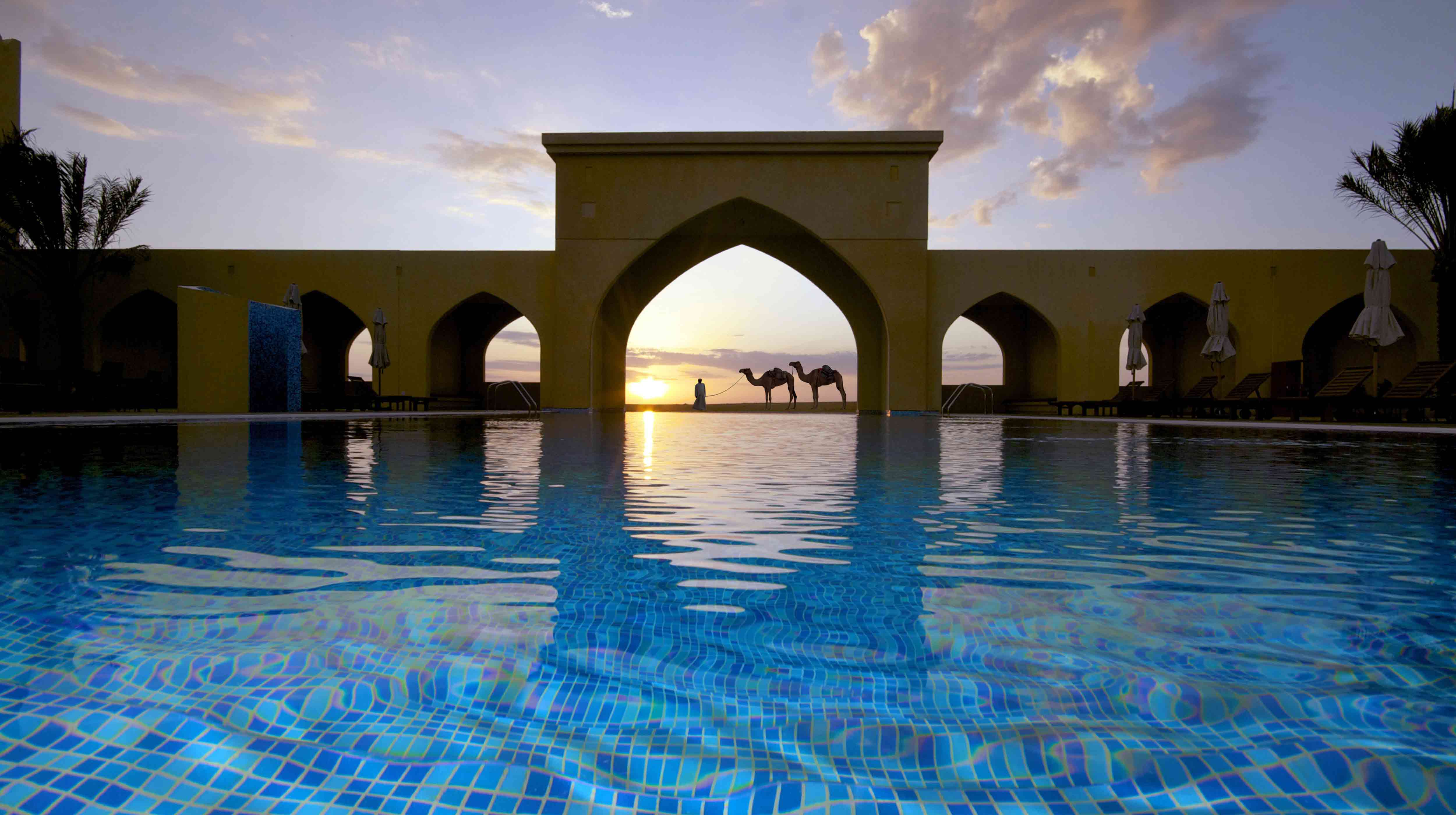 Man guiding two camels beyond the swimming pool at Tilal Liwa Hotel