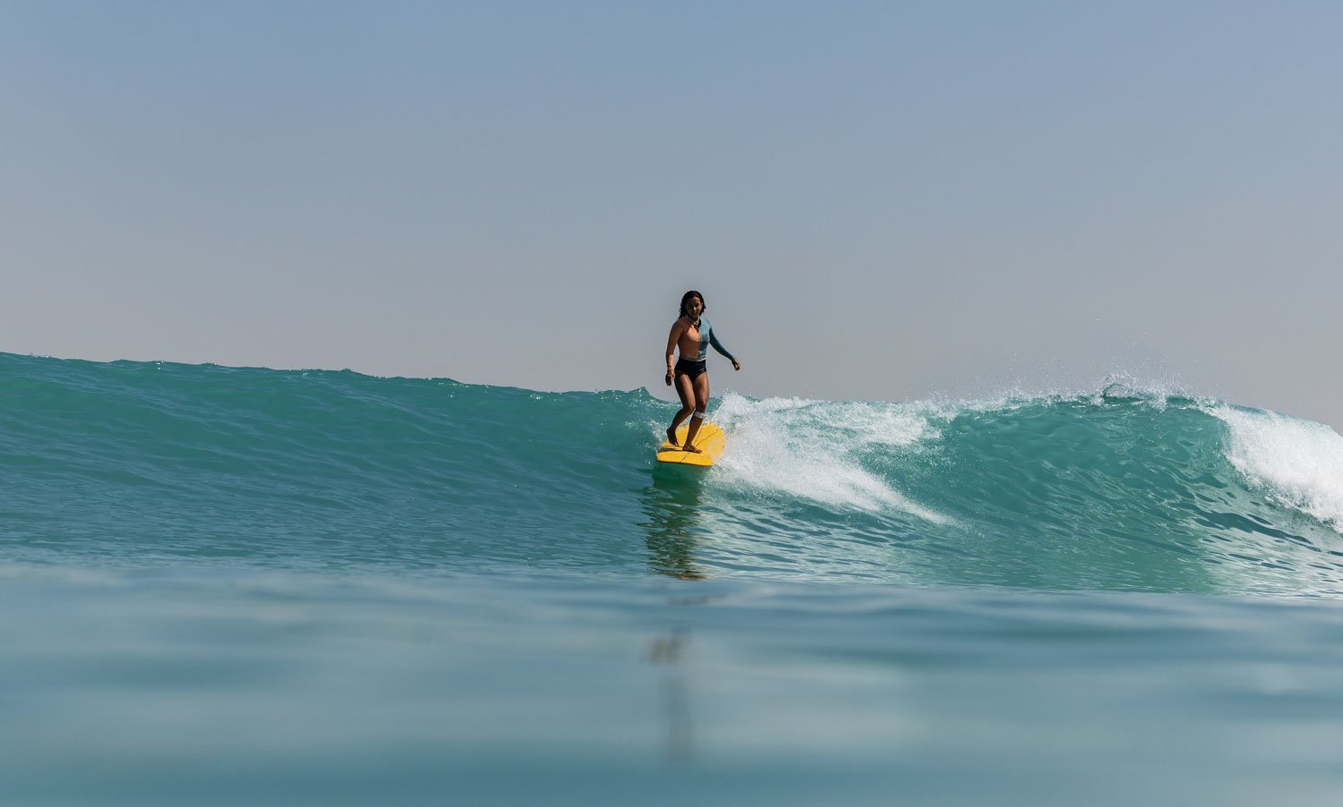 Woman surfing on a yellow board, riding a clear blue wave under a cloudless sky.
